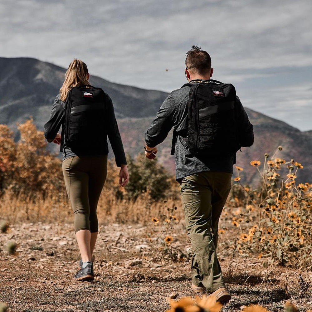 Two people with backpacks hike on a trail surrounded by wildflowers, with mountains in the background.