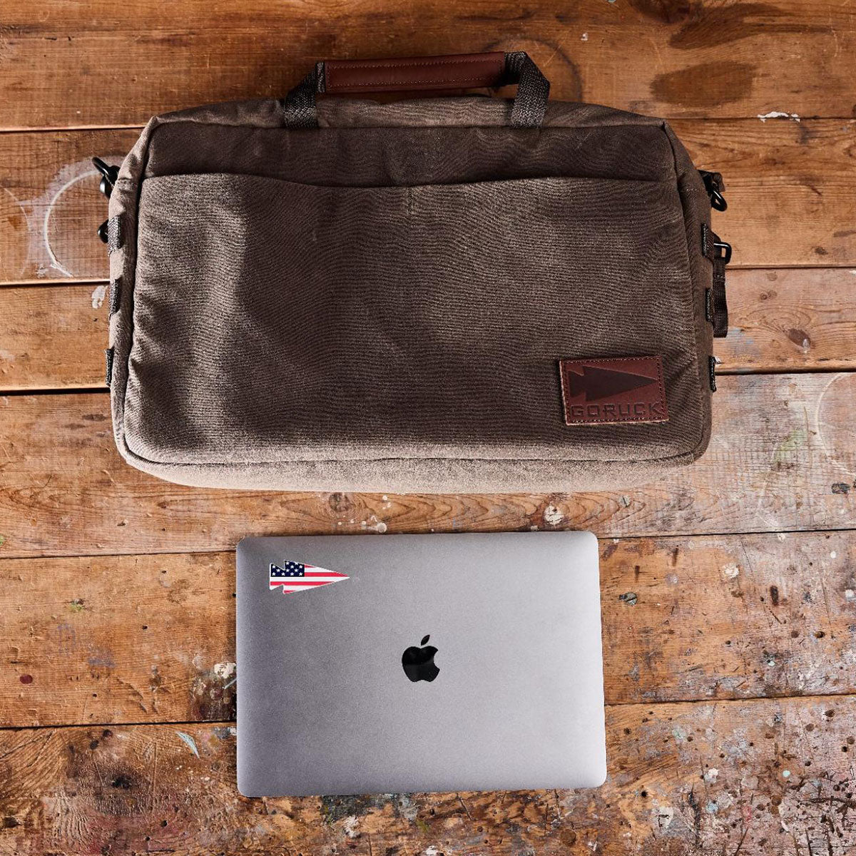 A brown GORUCK bag and a MacBook with a sticker on a scratched wooden surface.