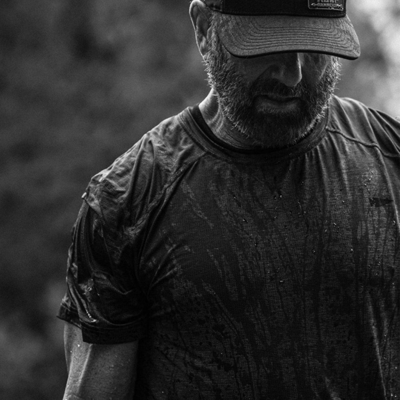 Bearded man in a hat and wet shirt standing outdoors, looking down, in black and white.