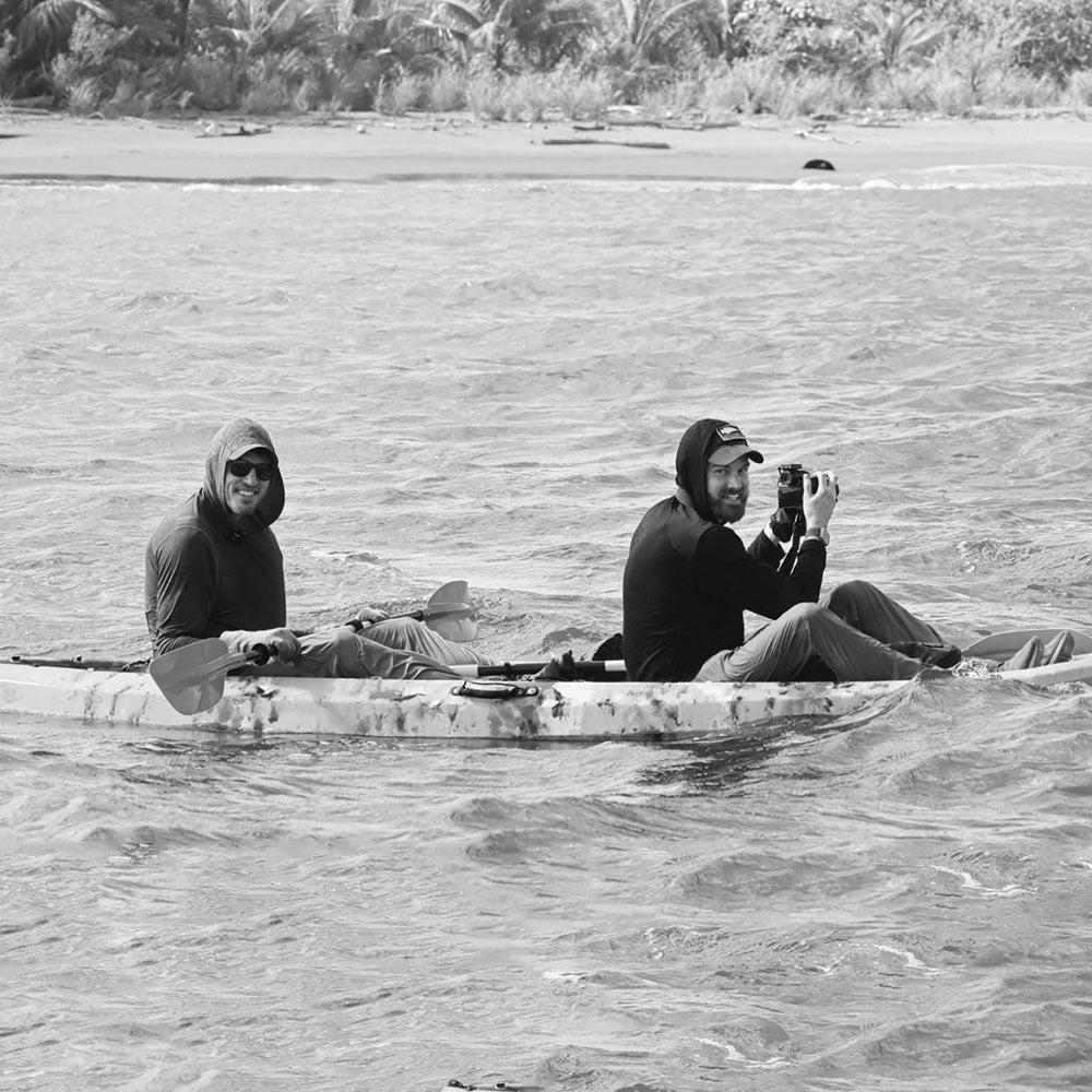 Two people in hoodies paddle a kayak; one smiles, the other holds a camera, on a body of water.