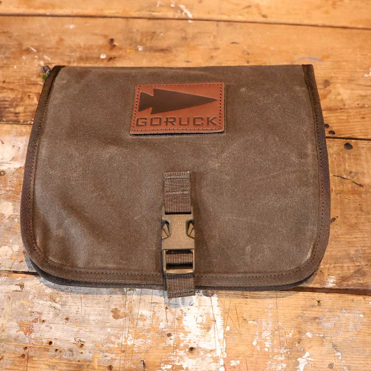 A brown GORUCK pouch with a buckle sits on a scratched wooden surface.