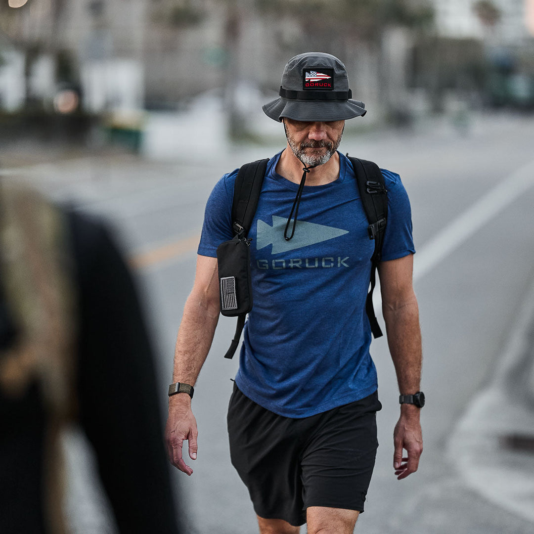 A man in a blue GORUCK shirt and black GORUCK Boonie Hat - Tactical - ToughDry with 50+ UPF walks down the street, carrying a backpack and water tube.