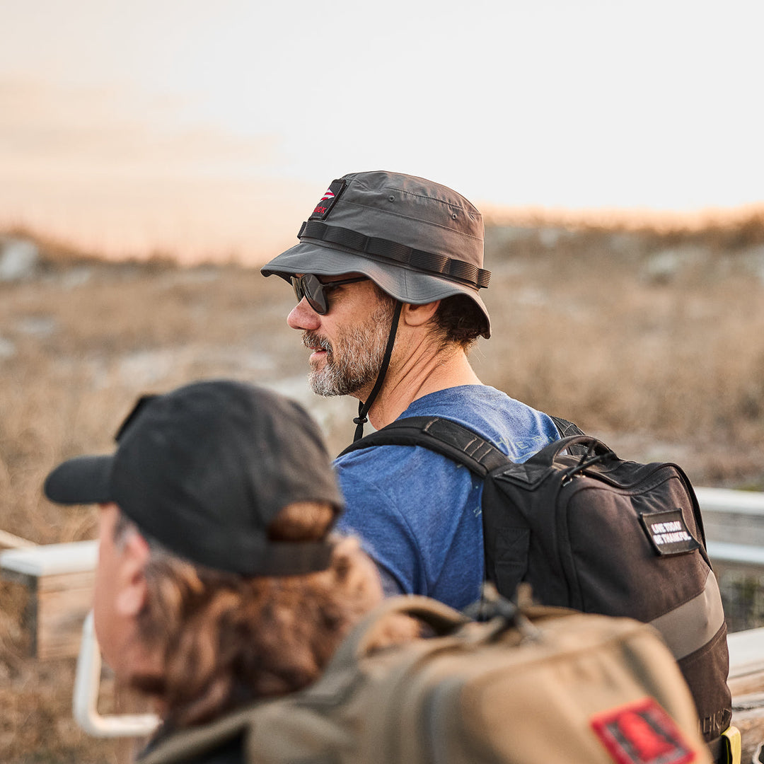 Two men wearing GORUCK Boonie Hat - Tactical - ToughDry backpacks hike in a dry, grassy landscape at sunset.