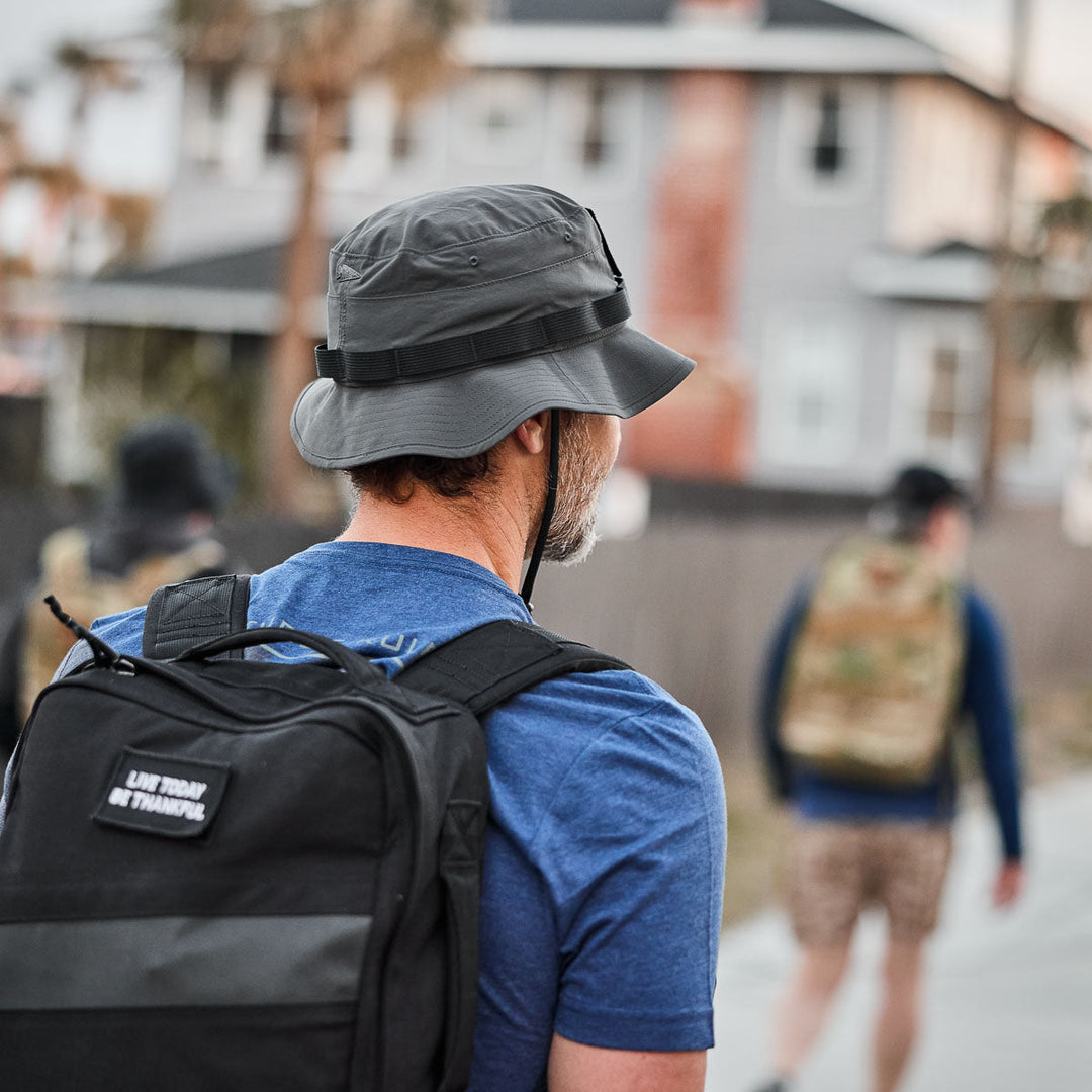 A man wearing a GORUCK Boonie Hat - Tactical - ToughDry in gray and a blue shirt with a black backpack walks outdoors, following others.