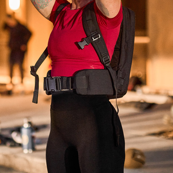 A person in a red shirt and black leggings, arms raised outdoors in soft light, wears a backpack with the Padded Hip Belt—ready for rucking or their next GORUCK event.