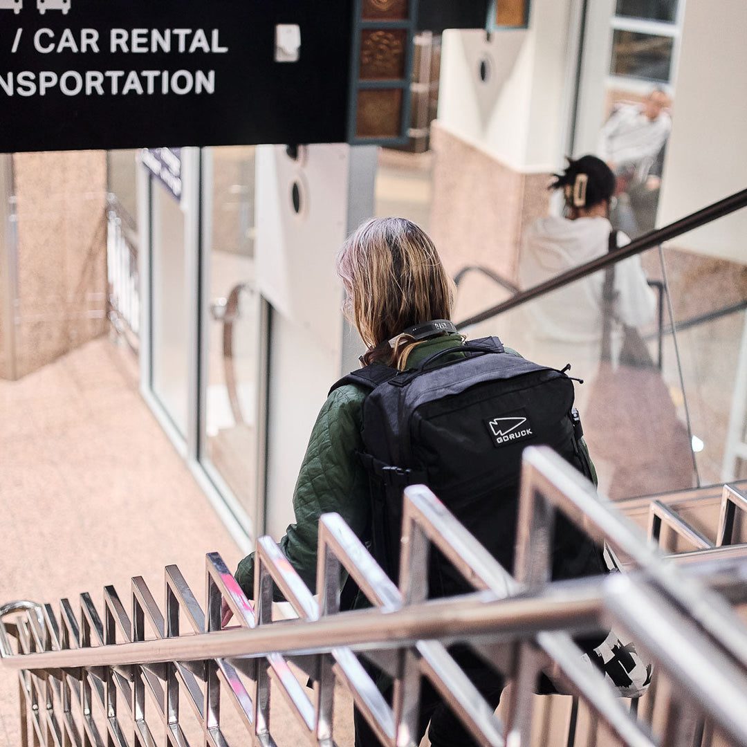 A person carrying a GORUCK GR3 - X-PAC waterproof backpack walks down an indoor staircase near a car rental and transportation sign.