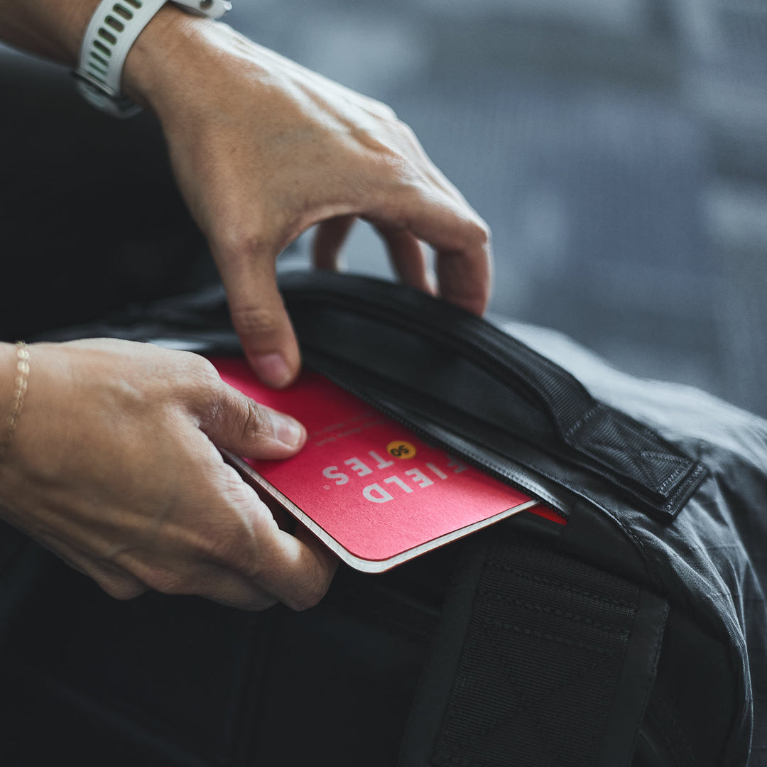 Hands placing a red FIELD NOTES notebook into a black GORUCK GR3 - X-PAC zippered bag made from waterproof material—ideal for carry-on use or your GR3 backpack.