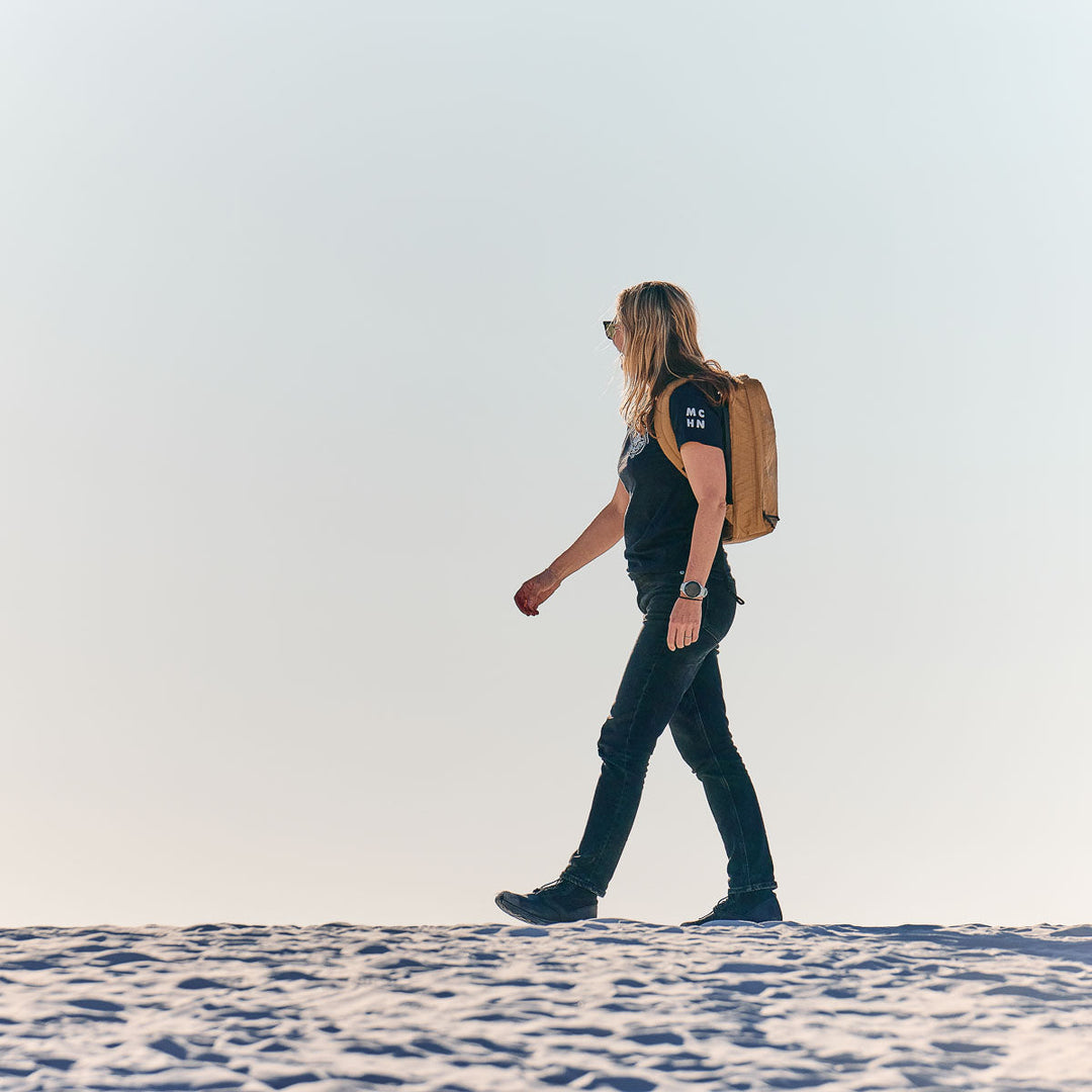 A person with long hair walks on sandy ground under a clear sky, wearing the lightweight, waterproof GORUCK GR0 - X-PAC backpack.