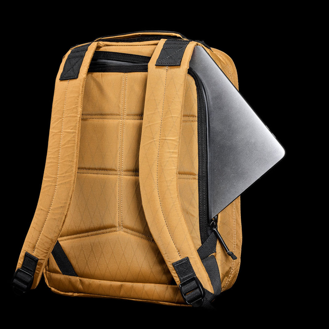 The GORUCK GR0 - X-PAC is a tan backpack with black straps, made from lightweight X-PAC fabric, shown with a silver laptop partially tucked into the back zipper pocket.