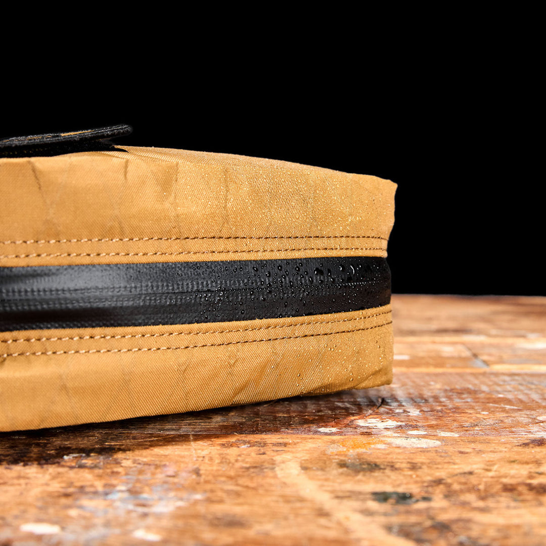 Close-up of the GORUCK GR2 Field Pocket - X-PAC, a yellow waterproof bag with a black zipper on a worn wood surface, water droplets visible—perfect for protecting camera gear.