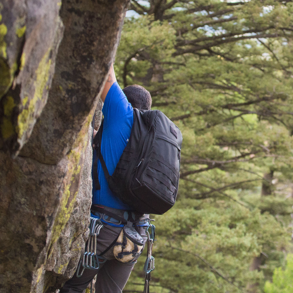 Wearing climbing gear and a GORUCK GR2 - USA ruck, a person scales a rock face amidst lush green trees.