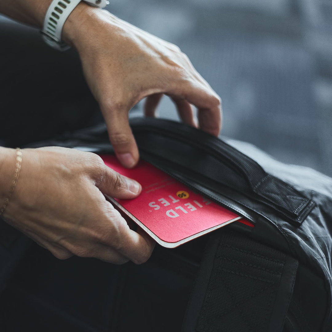 A person places a red Field Notes notebook into a black GORUCK GR3 - X-PAC carry-on backpack made from ultra-waterproof material.