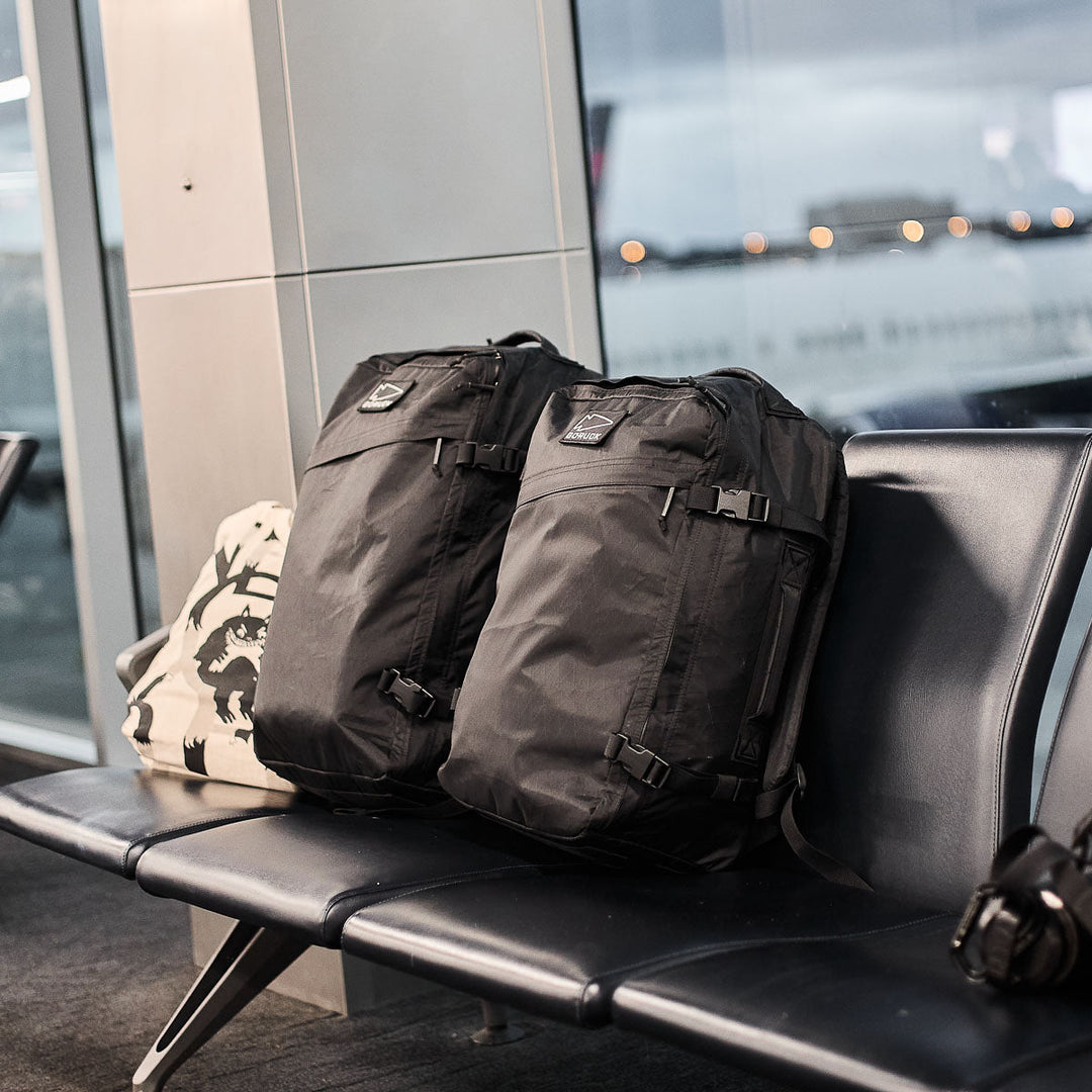Two black GORUCK GR3 - X-PAC ultra-waterproof backpacks and a white bag sit on airport chairs by a window with a plane outside.