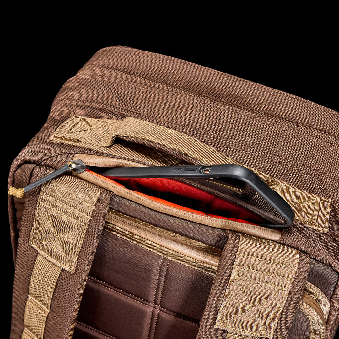 A GORUCK GR2 travel backpack in brown fabric features a smartphone partially sticking out of its zippered pocket.