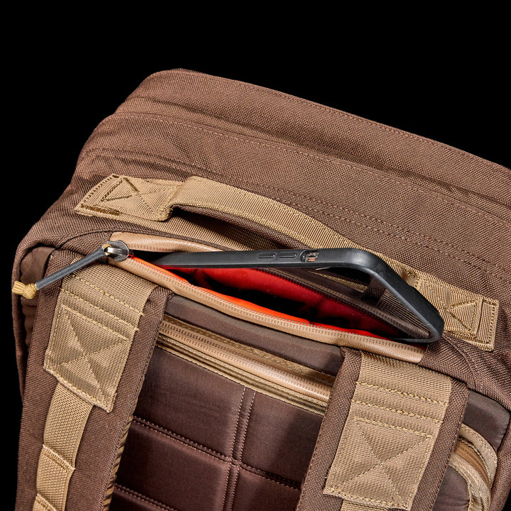 A GORUCK GR2 travel backpack in brown fabric features a smartphone partially sticking out of its zippered pocket.