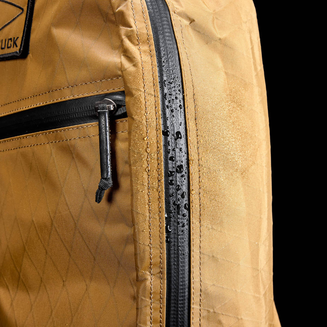 Close-up of the GORUCK Bullet X-PAC backpack in brown, made from weatherproof X-PAC material, with water droplets beading on its zipper and fabric.