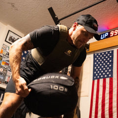 Joe lifts a 100 lb sandbag wearing the Achilles Vest, surrounded by an American flag and wall decorations.