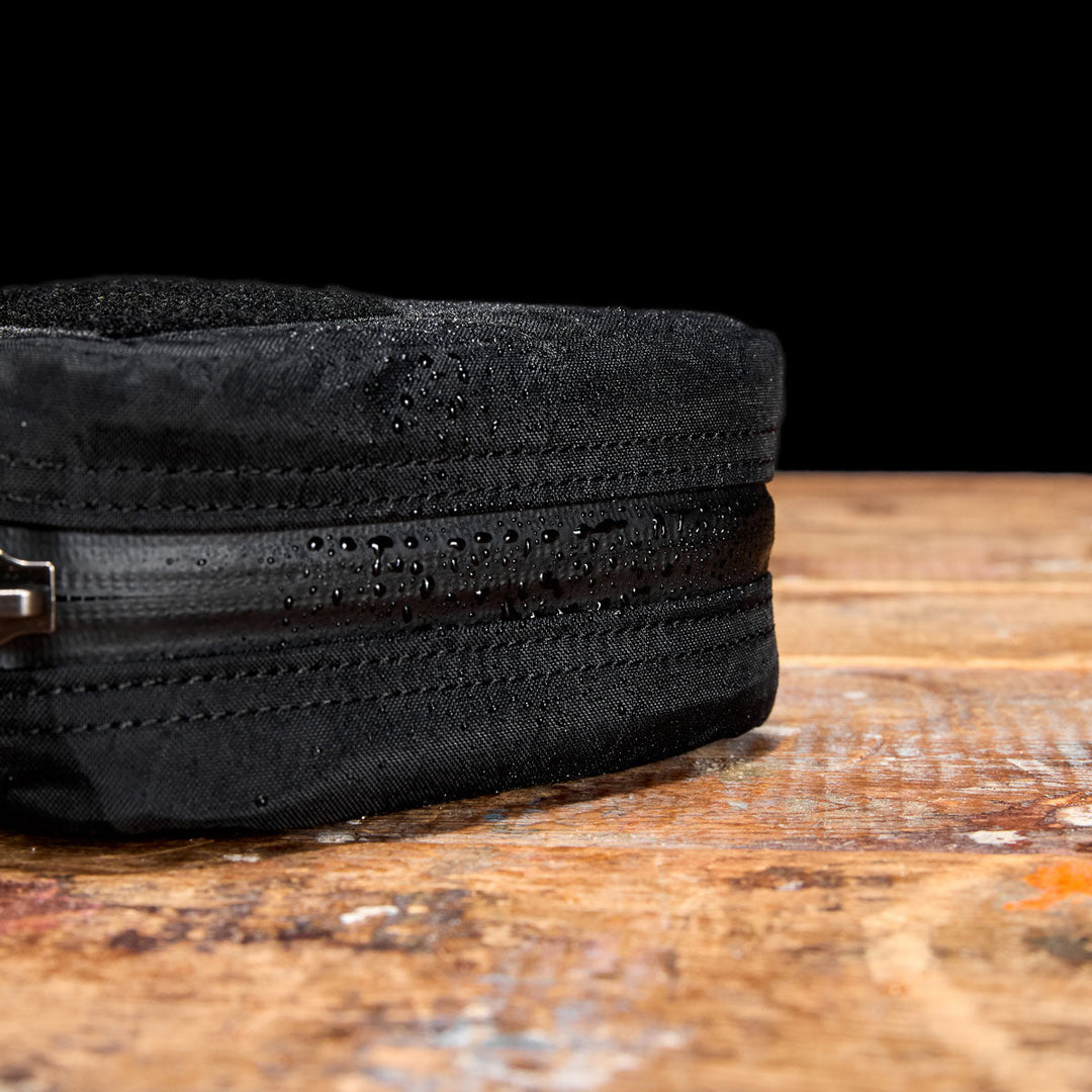 Close-up of the GORUCK GR1 Field Pocket - X-PAC, a black electronics organizer with waterproof fabric and AquaGuard zippers, shown with water droplets on its surface atop a wooden table.