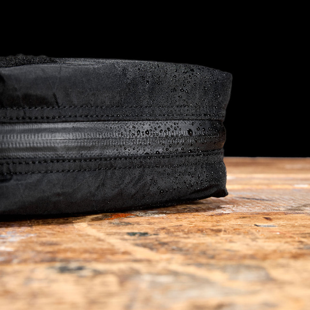 Close-up of the GORUCK GR2 Field Pocket - X-PAC, a black waterproof bag with water droplets on its zipper, resting on a wooden surface against a black background.