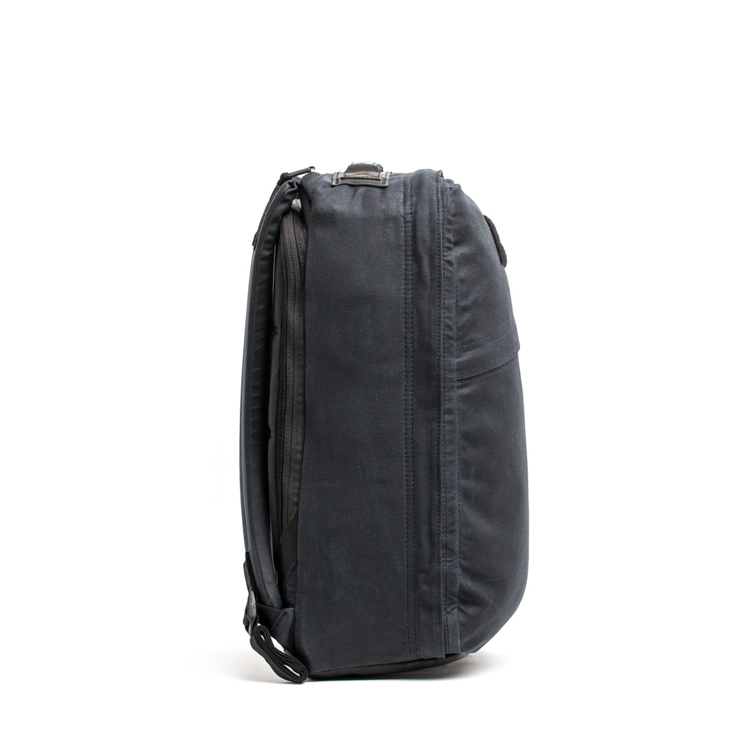 Side view of the closed GR1 USA - Heritage backpack in dark gray, made by GORUCK from durable waxed canvas, on a white background.