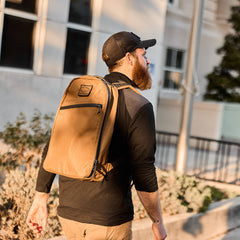 Man wearing tan GORUCK backpack and black cap outdoors, urban background