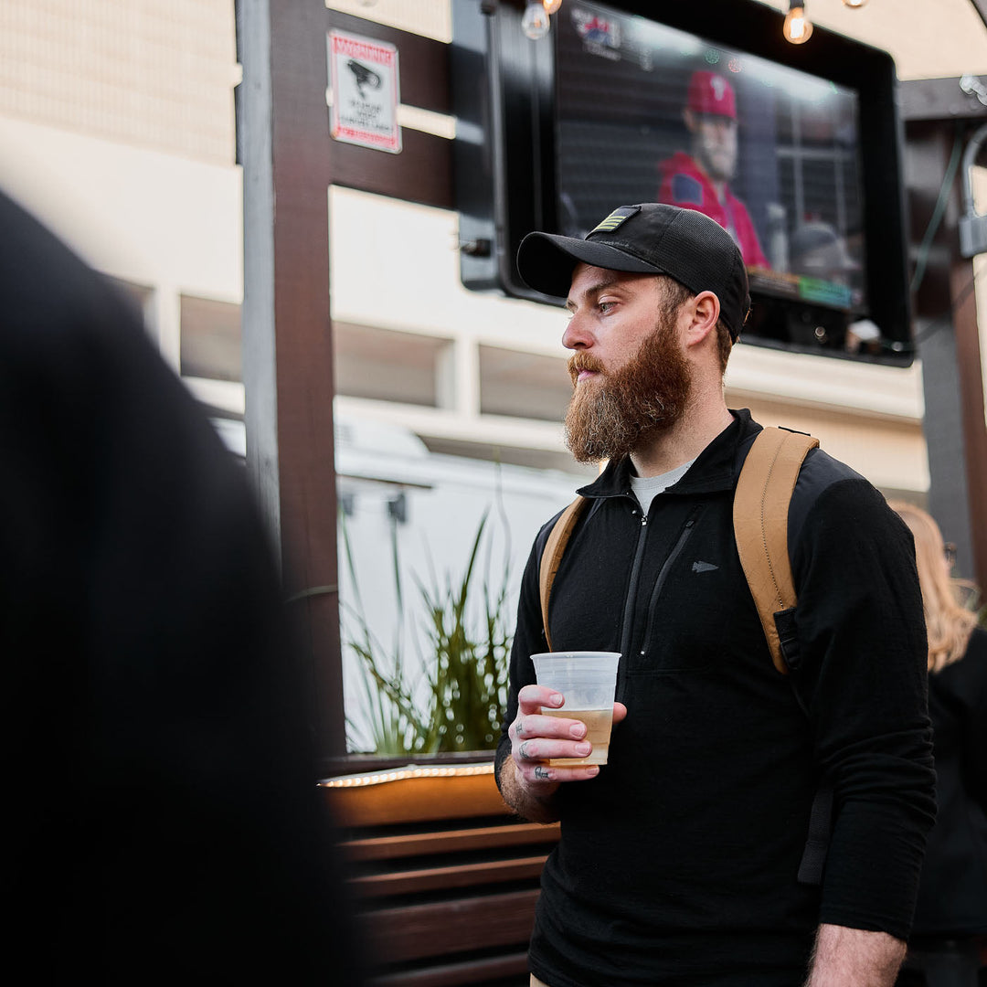 A bearded man with a cap carries the GORUCK Bullet X-PAC weatherproof backpack and holds a drink while standing outdoors near a TV displaying a baseball game.