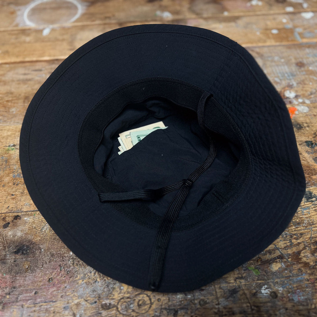 A GORUCK Boonie Hat - Slick - ToughDry rests on a wooden surface, with paper money tucked inside its inner band.