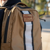 Close-up of a tan GORUCK EU Basic Rucker® backpack featuring a patch and vertical white and black stripe, ideal for carrying a ruck plate outdoors.