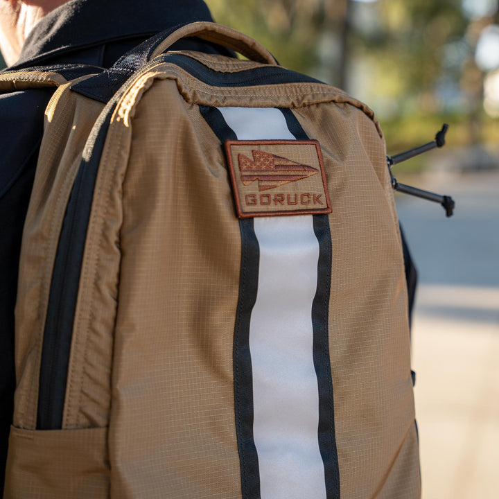Close-up of a tan GORUCK EU Basic Rucker® backpack featuring a patch and vertical white and black stripe, ideal for carrying a ruck plate outdoors.