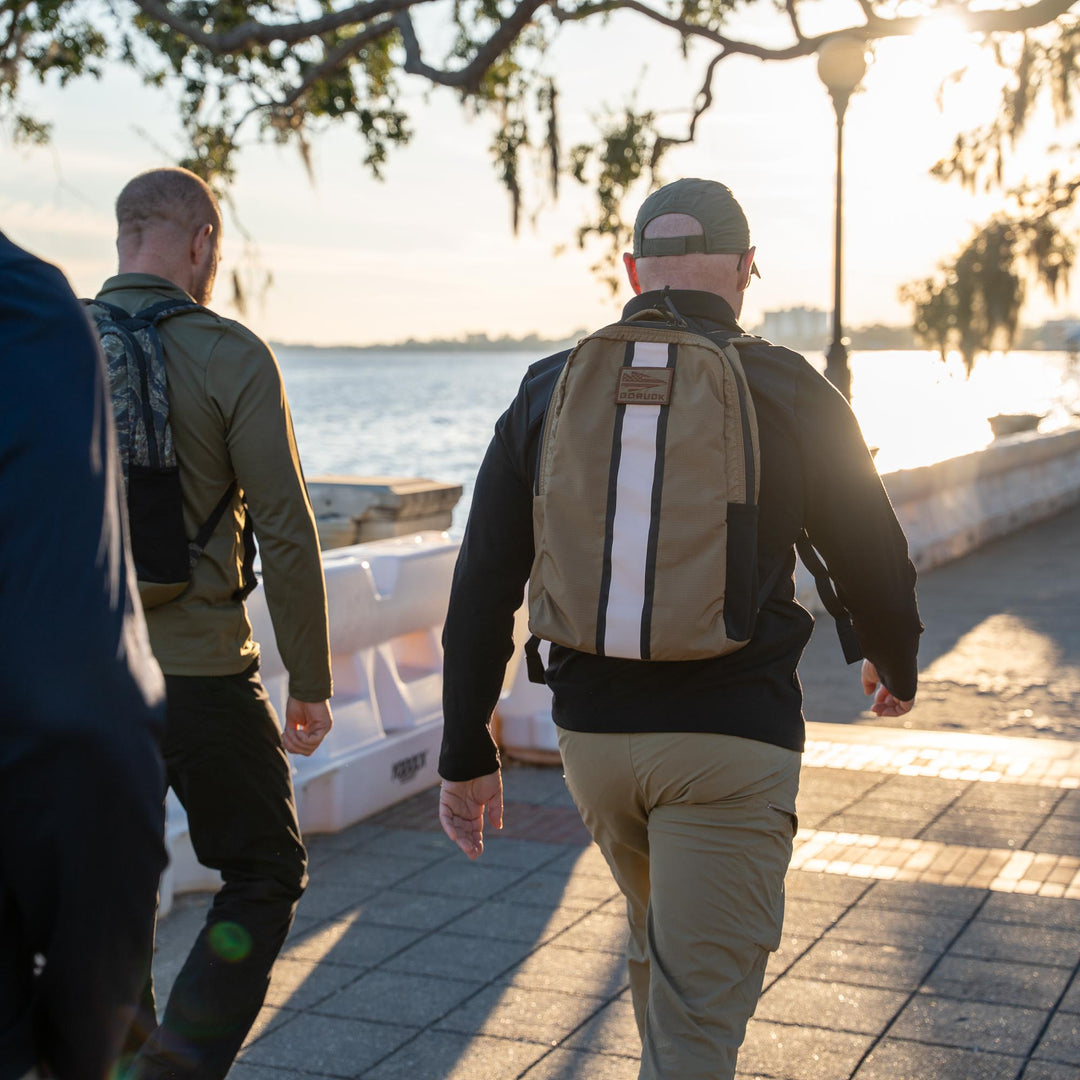 Two men with GORUCK EU Basic Rucker® backpacks walk along a waterfront path in the sunlight.