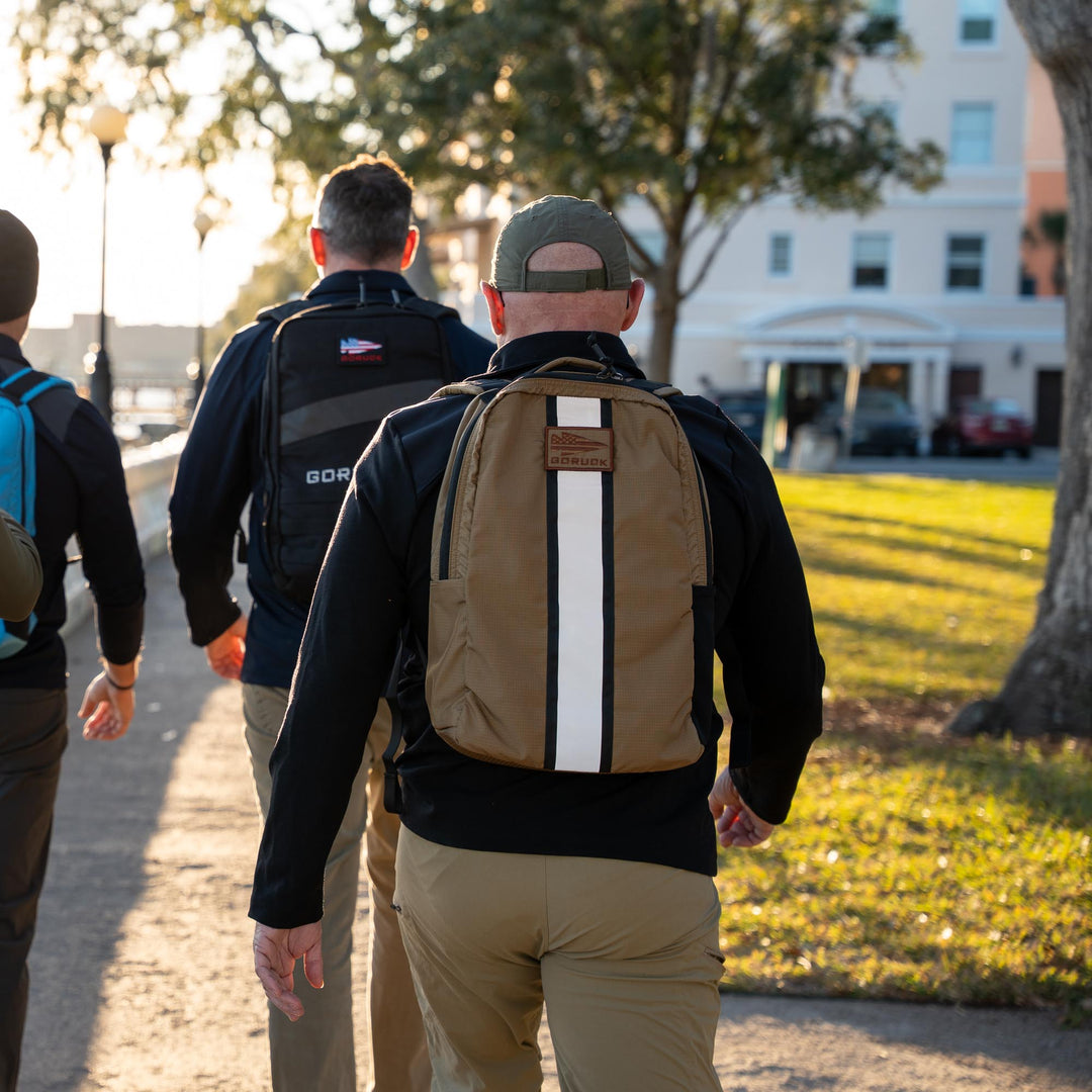 Three people carrying GORUCK EU Basic Rucker® backpacks walk along a sunlit path bordered by trees and a building.