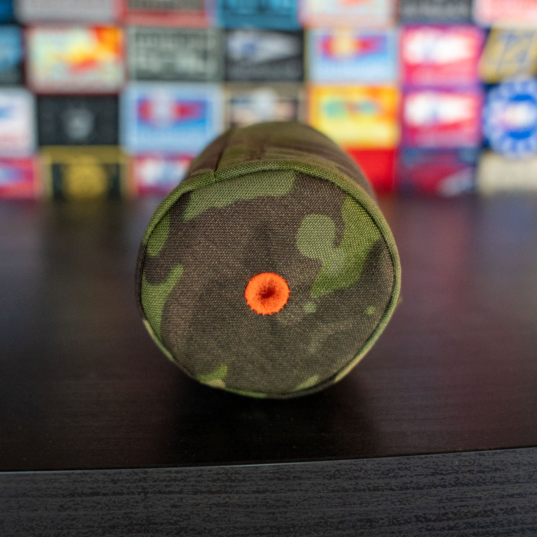 The GORUCK Ballistic Beer Jacket, made from tough 1000D CORDURA in a camouflage pattern with an orange grommet on the end, sits on a dark surface against a blurred colorful background.