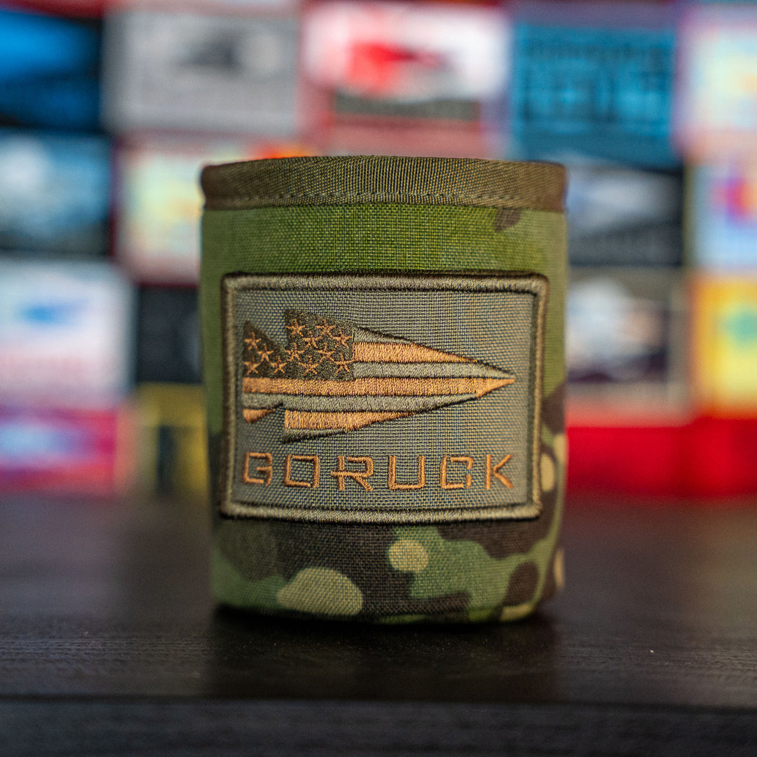 The Ballistic Beer Jacket - GORUCK by GORUCK is a camouflage beer koozie crafted from rugged 1000D CORDURA, featuring a GORUCK patch and colorful blurred patches in the background.