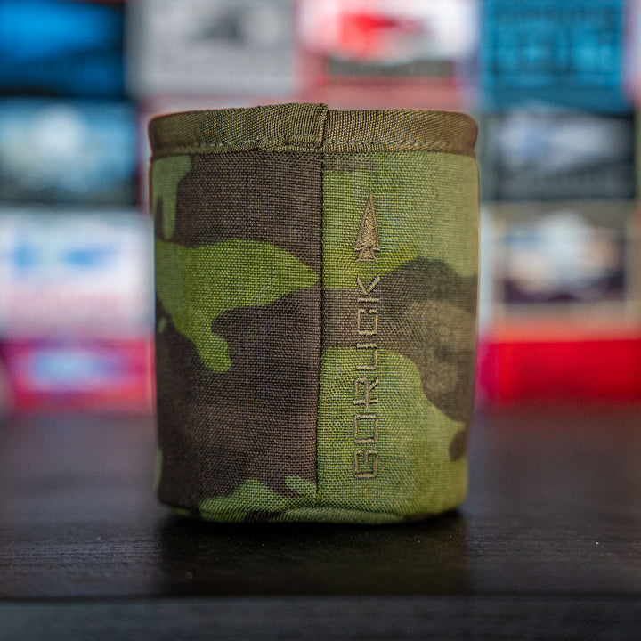 The Ballistic Beer Jacket by GORUCK is a camouflage-patterned 1000D CORDURA koozie with the "GORUCK" and arrow logo, displayed against a blurred, colorful background.