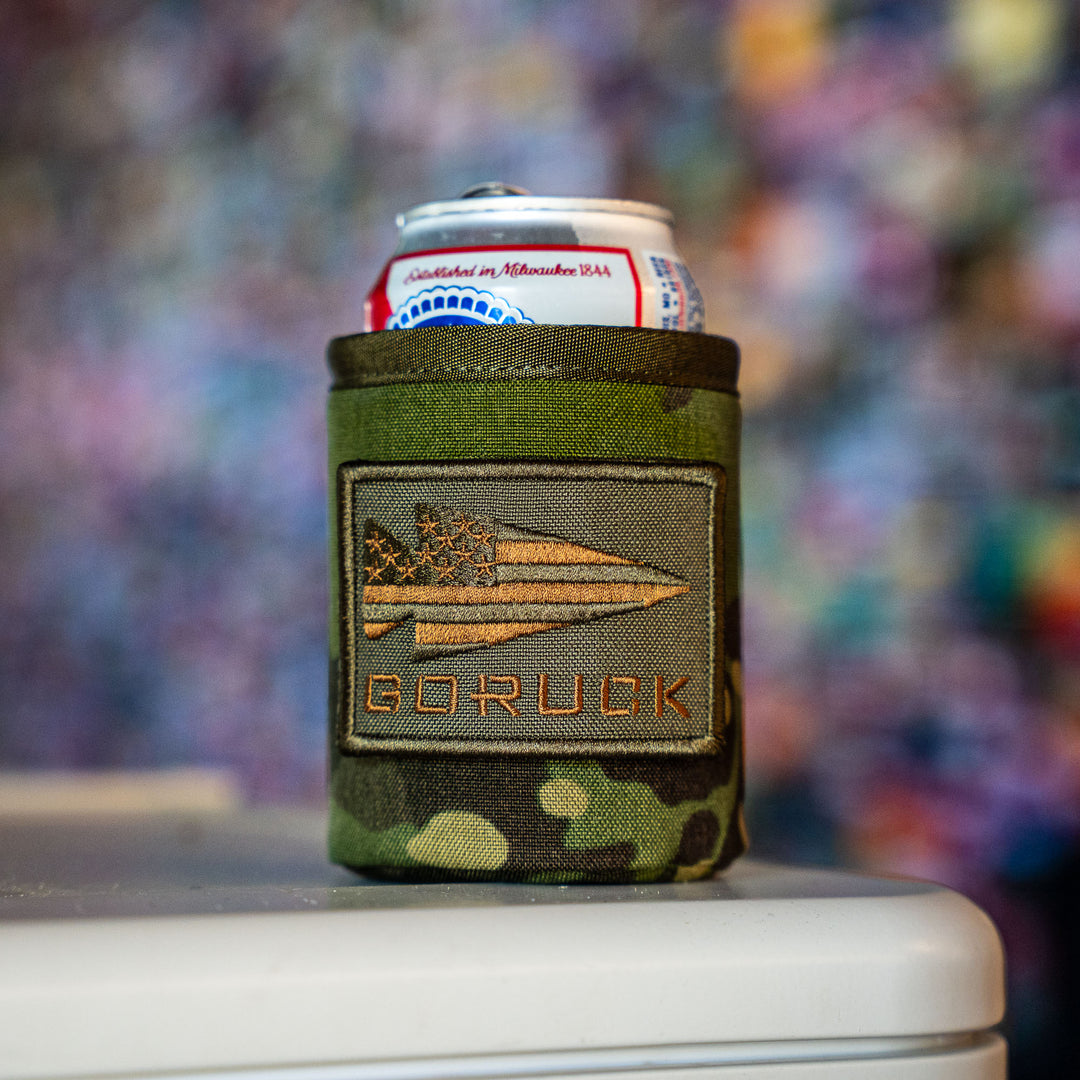 A can of beer sits on a white surface in the GORUCK Ballistic Beer Jacket, a rugged 1000D CORDURA koozie with a patch.