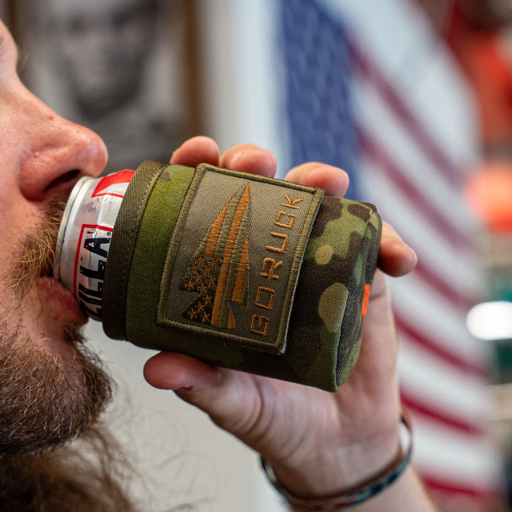 A person drinks from a can wrapped in the GORUCK Ballistic Beer Jacket, with a blurry American flag in the background.