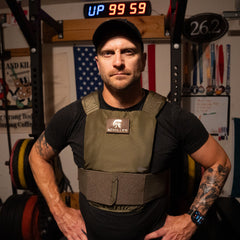A man in a cap and Achilles Vest stands in a gym with an American flag and weights behind him.