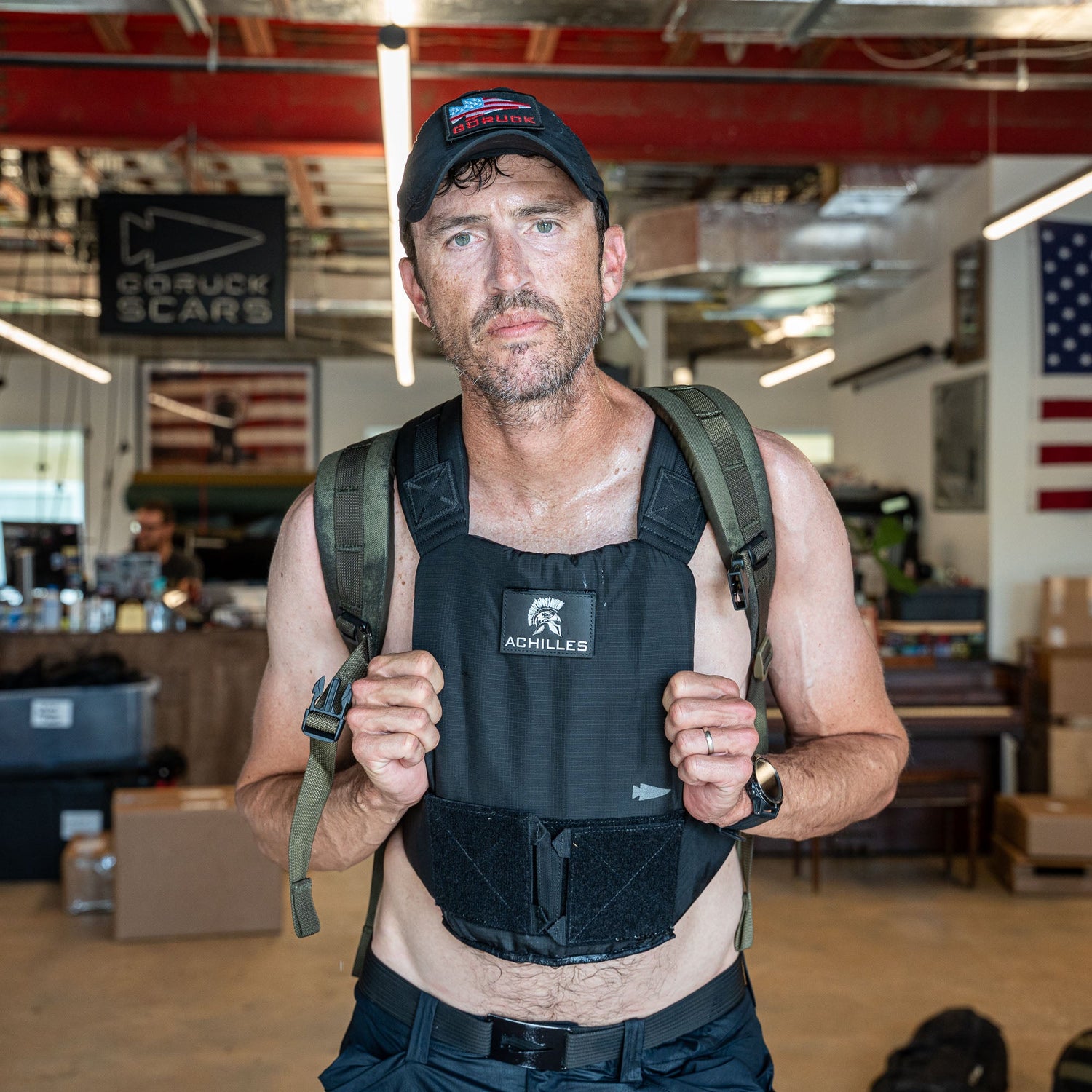 A man wearing the Achilles | Men's Weighted Vest, a cap, and a backpack stands indoors with American flags in the background.