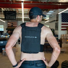 A man in a cap wears the Achilles | Men's Weighted Vest indoors, facing away, with an American flag on the wall.