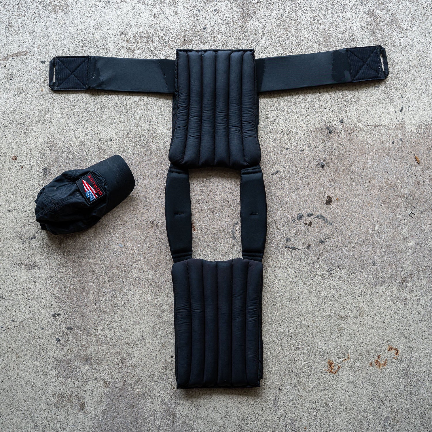 The Achilles | Men's Weighted Vest, featuring padded non-toxic construction, is laid flat on concrete beside a black baseball cap.
