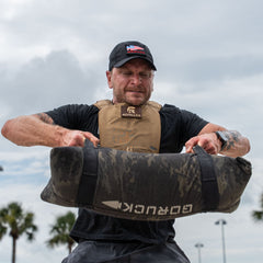 A man wearing the Achilles | Men's Weighted Vest lifts a heavy sandbag outdoors with palm trees and a cloudy sky in the background.