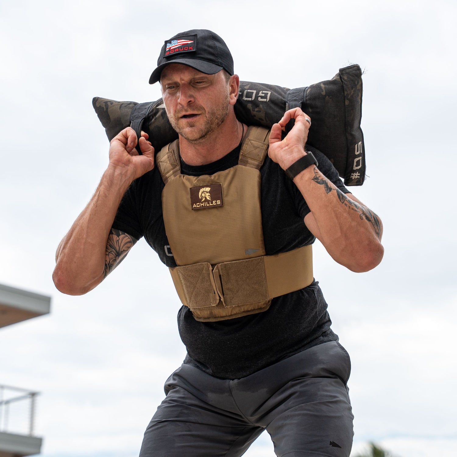 A man wearing the Achilles | Men's Weighted Vest lifts a 50lb sandbag on his shoulders during an outdoor workout.