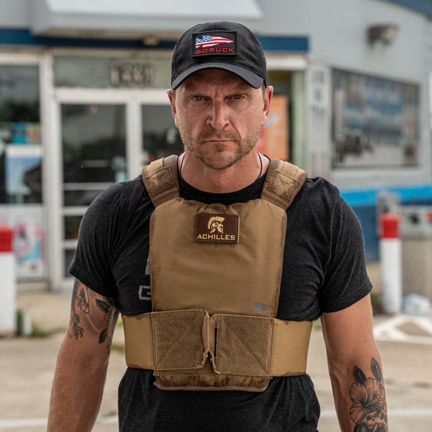 A man in a black shirt and cap wears the Achilles | Men's Weighted Vest in tan, standing outdoors in front of a building.