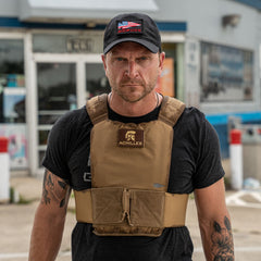 A man in a black shirt and cap wears the Achilles | Men's Weighted Vest in tan, standing outdoors in front of a building.