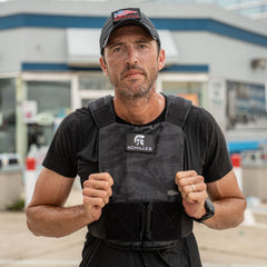A man stands outdoors wearing the Achilles | Men's Weighted Vest and a cap, looking at the camera with a building in the background.