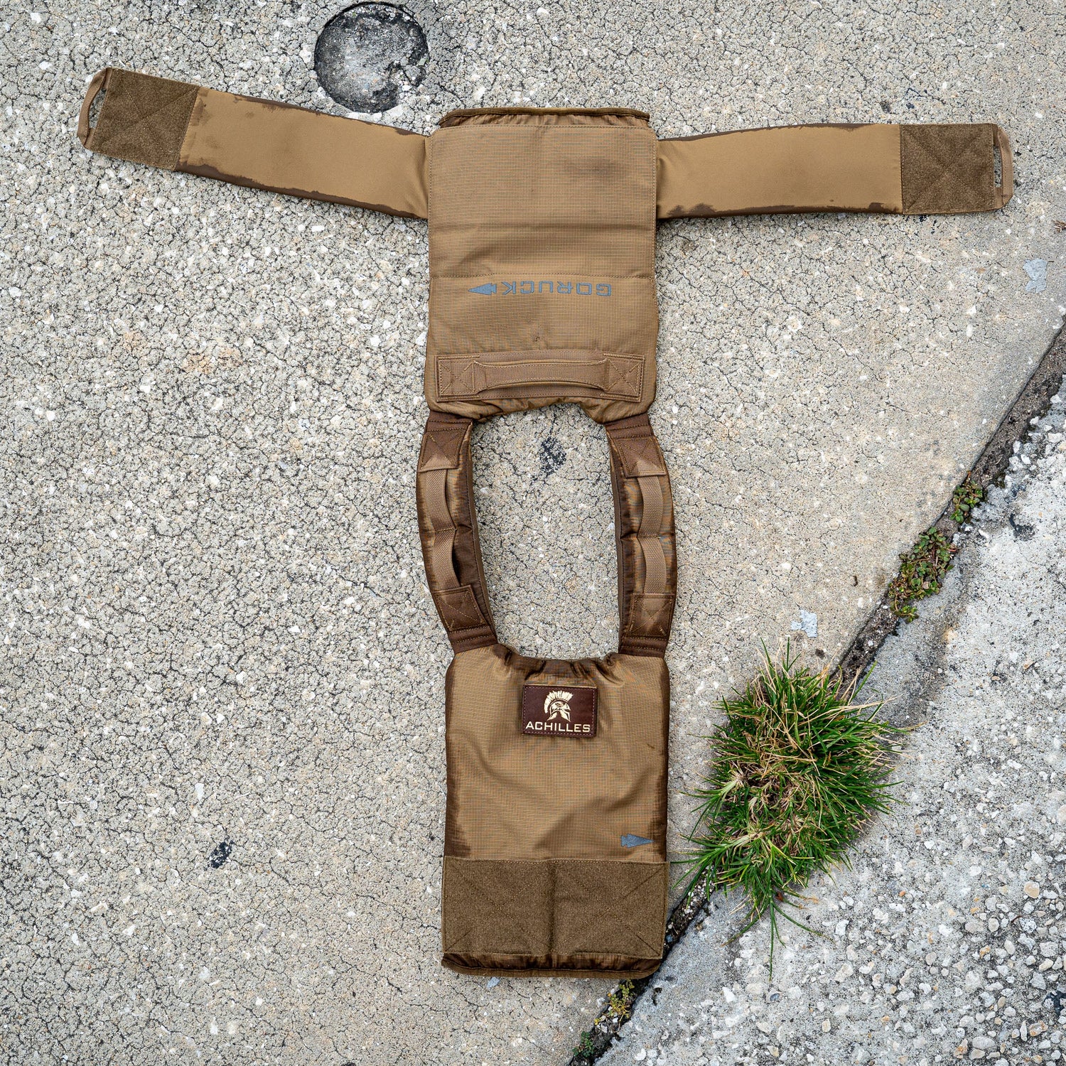 Two Achilles | Men's Weighted Vests in brown lay open on cracked concrete beside a small patch of grass.