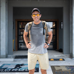 A man in sunglasses and a gray t-shirt stands outside a building with a backpack, sweating visibly while wearing GORUCK Men’s Training Shorts – ToughStretch.