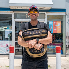 Smiling man in sunglasses and hat holds a GORUCK Sand Tombstones portable workout sandbag outside a store on a sunny day.