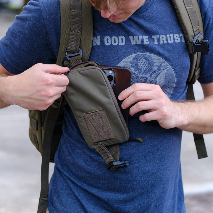 Wearing a blue t-shirt, a person places their phone into the GORUCK Sternum Pouch - Cordura, attached via MOLLE to their backpack strap.