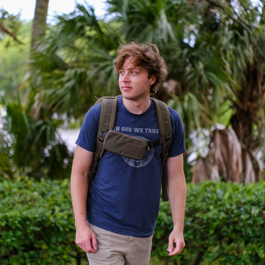 A young man stands outdoors amid greenery, wearing a blue shirt and khaki pants, with a GORUCK Sternum Pouch - Cordura attached to his backpack.